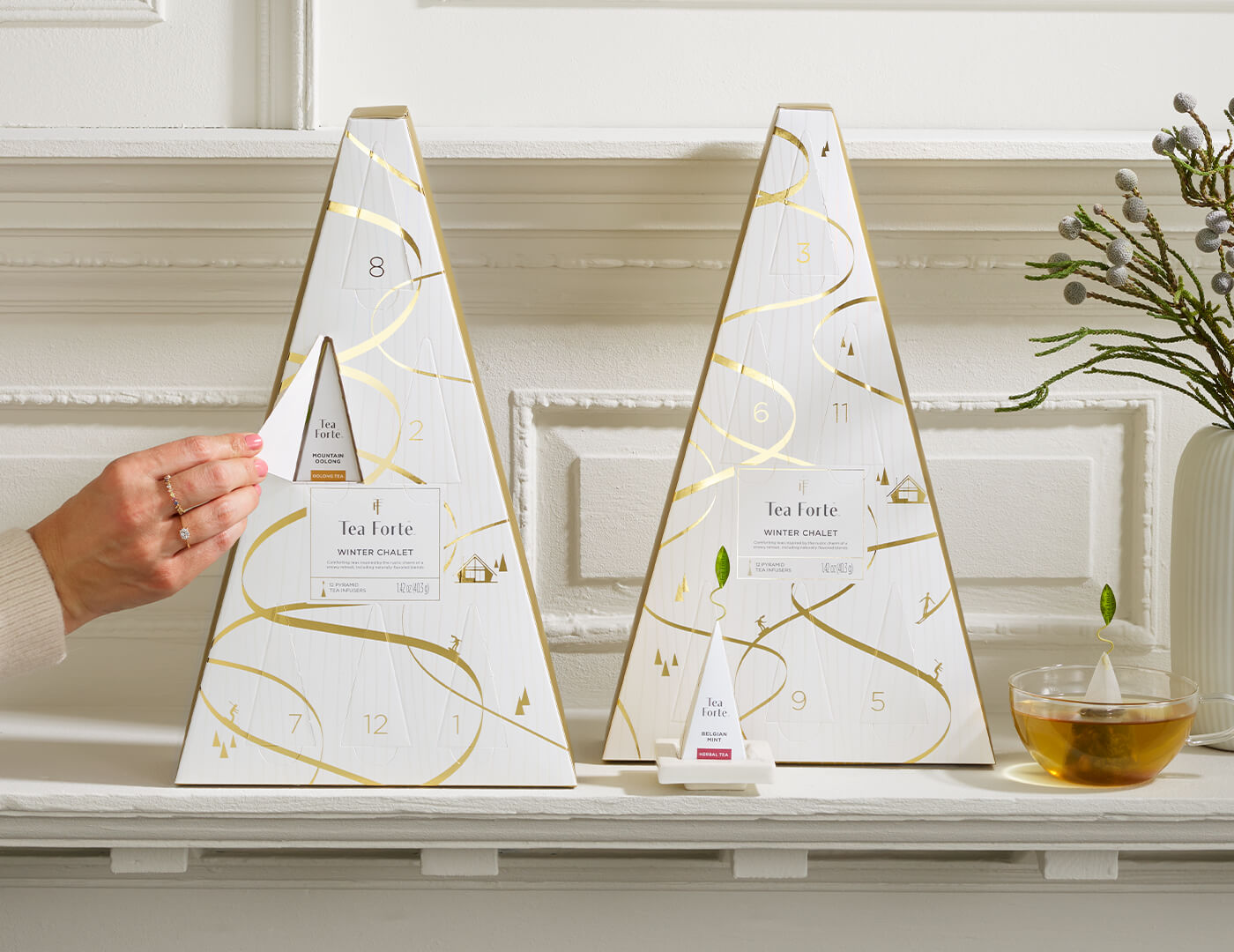 Winter Chalet Advents, one with doors closed, the other being opened with a female hand, on a white shelf with glass teacup of tea